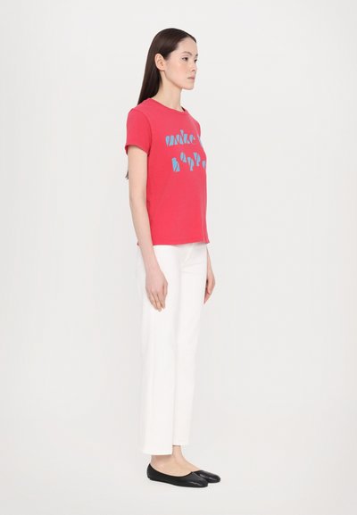 Red cotton t-shirt with blue graphic design, paired with white ankle-length pants and black flat shoes. Simple, fitted design, short sleeves.
