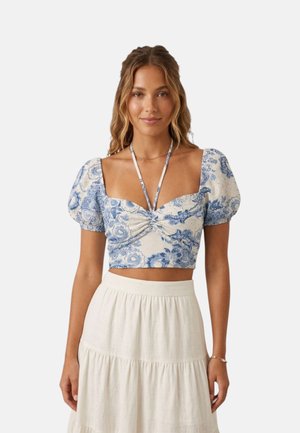 Donna che indossa un top corto floreale blu e bianco con maniche a sbuffo e una gonna bianca a vita alta, in piedi davanti a uno sfondo semplice.