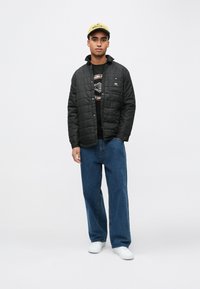 Czarna pikowana kurtka na suwak, noszona na czarnej koszulce z nadrukiem, zestawiona z luźnymi niebieskimi dżinsami i białymi trampkami.