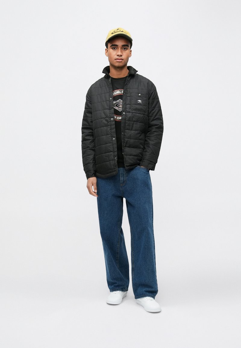 Czarna pikowana kurtka na suwak, noszona na czarnej koszulce z nadrukiem, zestawiona z luźnymi niebieskimi dżinsami i białymi trampkami.