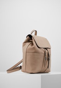 Beige läderryggsäck med klaffstängning, justerbara remmar, en framficka och svarta metalldetaljer. Slät yta och minimalistisk design.