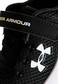Zapatilla deportiva negra con parte superior de malla, puntera reforzada y correa elástica. Presenta un logo blanco, diseño texturizado y sistema de cordones tradicional.