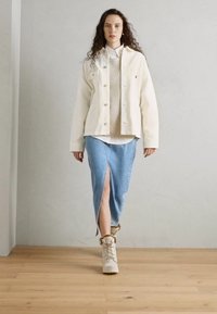Hvit oversized denimjakke over en beige strikkegenser, kombinert med et langt lys blått denimskjørt med splitt foran og kremfargede støvler.
