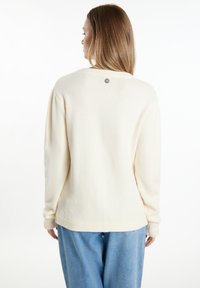 Crème-kleurige trui met een zachte textuur, lange mouwen en een ronde halslijn. Heeft een klein logo op de rug bij de hals. Gekleed met blauwe broek.