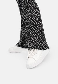 Weiße Sneakers mit flachen Sohlen und Schnürsenkeln, kombiniert mit schwarzen Schlaghosen mit einem weißen Blumenmuster auf schwarzem Hintergrund.