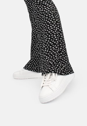 Sneakers bianchi con suola piatta e lacci, abbinati a pantaloni neri a palazzo con un motivo floreale bianco su sfondo nero.
