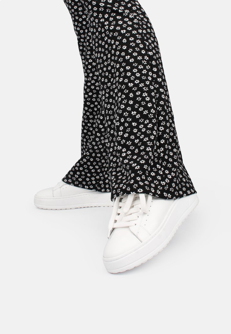 Weiße Sneakers mit flachen Sohlen und Schnürsenkeln, kombiniert mit schwarzen Schlaghosen mit einem weißen Blumenmuster auf schwarzem Hintergrund.