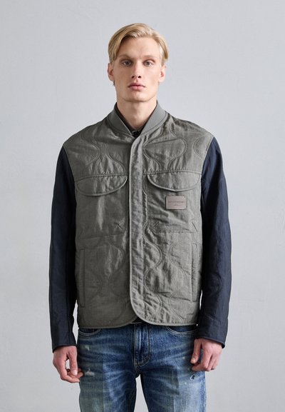 Gilet trapuntato grigio con texture liscia, due tasche sul petto e toppa con logo, indossato sopra una maglia a maniche lunghe scura e jeans di denim blu.