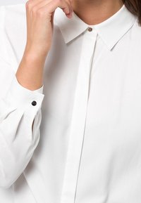 Witte blouse met een klassieke kraag, knopenlijst en geplooide mouwen. Voorzien van een kleine zwarte knoop op de manchette. Glad textuur van de stof.