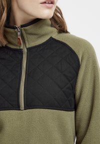 Pullover in pile verde oliva con un pannello frontale trapuntato nero. Presenta un colletto con zip e tessuto testurizzato.