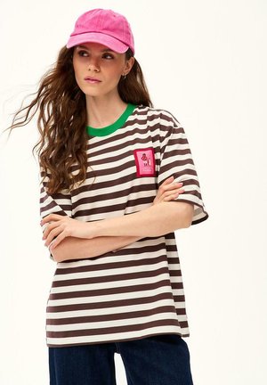 Kobieta z długimi brązowymi włosami, nosząca różową czapkę, brązowo-białą koszulkę w paski z zielonym kołnierzykiem i naszywką, z założonymi rękami.