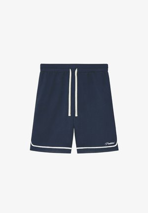 Mężczyzna w granatowych siateczkowych szortach z beżowym obszyciem, białych skarpetkach z logo oraz beżowych zamszowych sznurowanych butach, stojący na jednolitym tle.