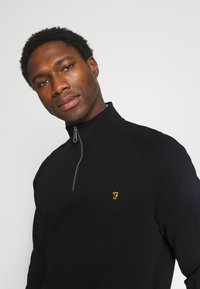 Black zip-up fleece with a high collar, featuring an orange embroidered logo on the chest and contrasting stitching details.