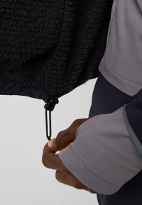 Fleecejacke mit einem strukturierten schwarzen Innenfutter, grauen Ärmeln und einem verstellbaren schwarzen Kordelzug am unteren Rand für eine eng anliegende Passform.