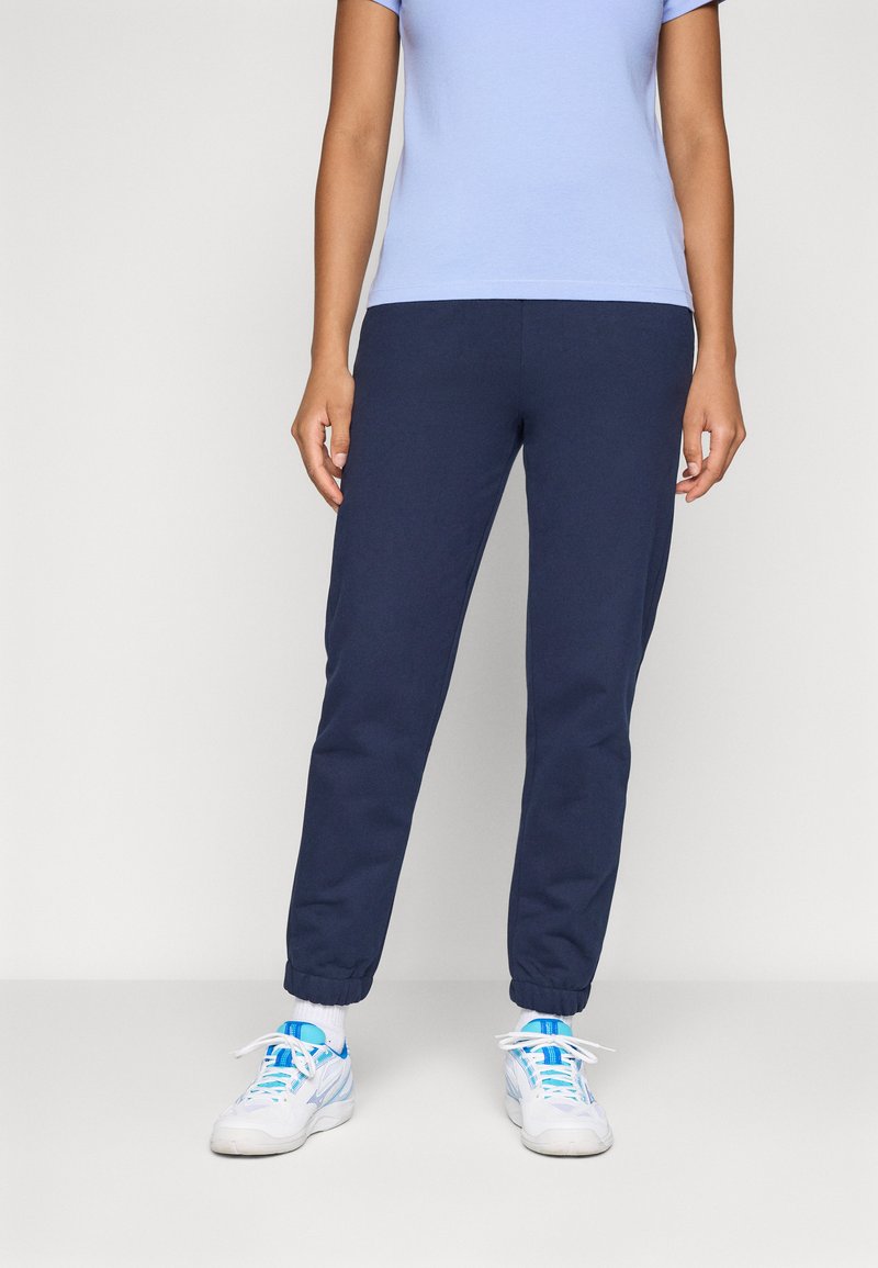 Person som har på seg marineblå joggebukser, hvite sneakers med blå detaljer, hvite sokker og en lys lilla t-skjorte med korte ermer, som står mot en ensfarget bakgrunn.