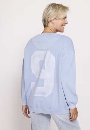 Blonde Frau lächelt, trägt ein hellblaues übergroßes Sweatshirt mit großer weißer Nummer 9 auf dem Rücken, kombiniert mit hellgrauen Hosen.