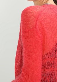 Gestrickter Pullover in leuchtendem Koralle, mit einer lockeren, offenen Maschenstruktur und durchsichtigen Abschnitten an Ärmeln und Rücken, die eine strukturierte, weiche Optik bieten.