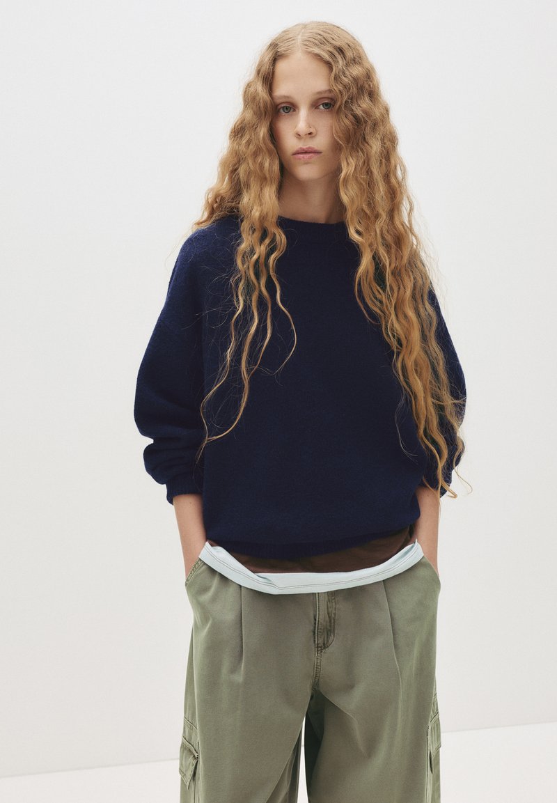 Ung kvinde med langt bølget blondt hår iført en mørk marineblå sweater og løse grønne cargobukser med hænderne i lommerne, stående mod en ensfarvet baggrund.