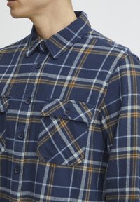 Camisa de franela de cuadros azul con líneas contrastantes en naranja y blanco, dos bolsillos frontales y cierres de botón. Textura suave con cuello tipo camisa.
