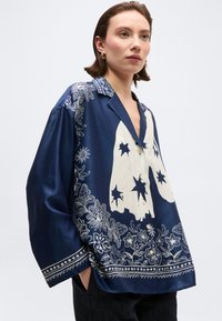 Γυναίκα που φοράει μια άνετη μπλε ναυτική πουκάμισα με κουμπιά, με μεγάλα λευκά σχέδια φαντασμάτων και αστεριών και λουλουδένια μοτίβα κατά μήκος του ποδόγυρου και των μανικιών.