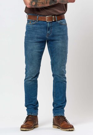 Férfi kék slim-fit farmernadrágban, barna bőrövvel és barna fűzős csizmával, karját keresztbe téve áll.