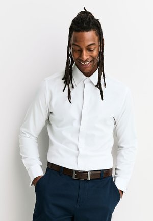 Man met dreadlocks, gekleed in een wit overhemd met knopen, een donkere riem en donkerblauwe broek, glimlacht en kijkt naar beneden met zijn handen in de zakken.