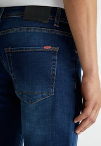 Donkerblauwe denim jeans met oranje stiksels, een leren zwarte tailleband en een klein rood merkembleem op de achterzak. Slim fit ontwerp.