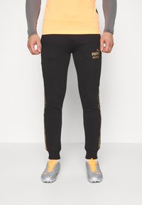 Černé sportovní kalhoty se zlatým označením po straně, tapered design a žebrované lemy, přidané se šedými fotbalovými kopačkami a oranžovými tkaničkami.