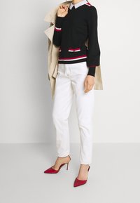 Schwarzer Pullover mit roten und weißen Streifen, weißem Kragen, kombiniert mit weißen Jeans und roten Pumps mit spitz zulaufender Spitze. Akzenttaschen mit Knöpfen.