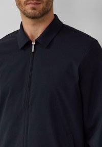 Marineblauwe jas met een kraag en een ritssluiting aan de voorkant. Zacht materiaal met een gladde textuur en zijzakken. Eenvoudig ontwerp met strakke lijnen.
