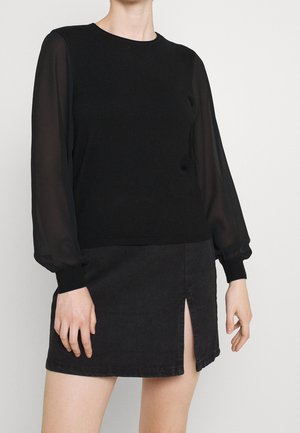 Personne portant un haut noir à manches longues transparentes et une mini-jupe noire fendue devant, debout devant un fond uni.
