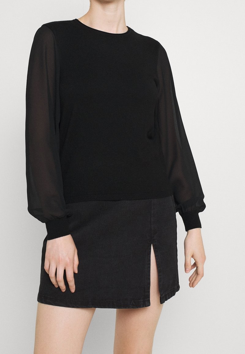 Personne portant un haut noir à manches longues transparentes et une mini-jupe noire fendue devant, debout devant un fond uni.