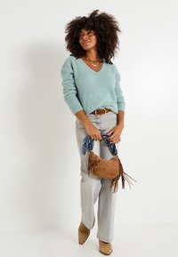 Suéter de pelaje azul claro con escote en V, combinado con jeans acampanados gris claro. Sosteniendo un bolso de ante marrón con acentos de bufanda azul y flecos.
