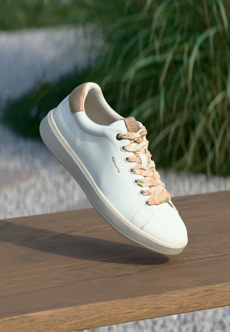 Λευκό δερμάτινο sneaker με υφασμάτινη τακτική τακούνι σε καφέ απόχρωση, κορδόνια με ροζ και πράσινο μοτίβο και μαλακή λάστιχη σόλα, να αιωρείται πάνω από ξύλινη επιφάνεια.