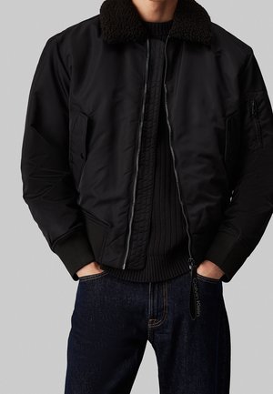 Hombre con chaqueta bomber negra con cuello de forro polar y puños acanalados, suéter de punto oscuro y vaqueros azules oscuros sobre fondo gris.