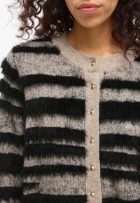 Cardigã bege com riscas felpudas pretas, apresentando um decote redondo e botões dourados à frente. Material de malha texturizada.