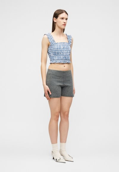 Femme debout, portant un haut court sans manches à carreaux bleus et blancs, un short taille haute gris, des chaussettes blanches et des chaussures blanches à bouts pointus.