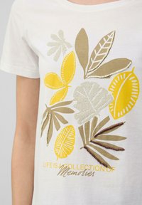 T-shirt en coton blanc avec un imprimé de feuilles jaunes et beiges aux accents texturés. Le texte indique "LA VIE EST UNE COLLECTION DE Souvenirs" en gris.