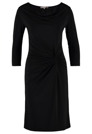 Femme marchant portant une robe noire longueur genou, des collants noirs transparents, des bottines noires et tenant une pochette beige.