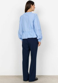 Donna con i capelli lunghi e scuri, vestita con una blusa leggera azzurro chiaro a balze e pantaloni larghi blu navy, in piedi di fronte a un muro bianco semplice.
