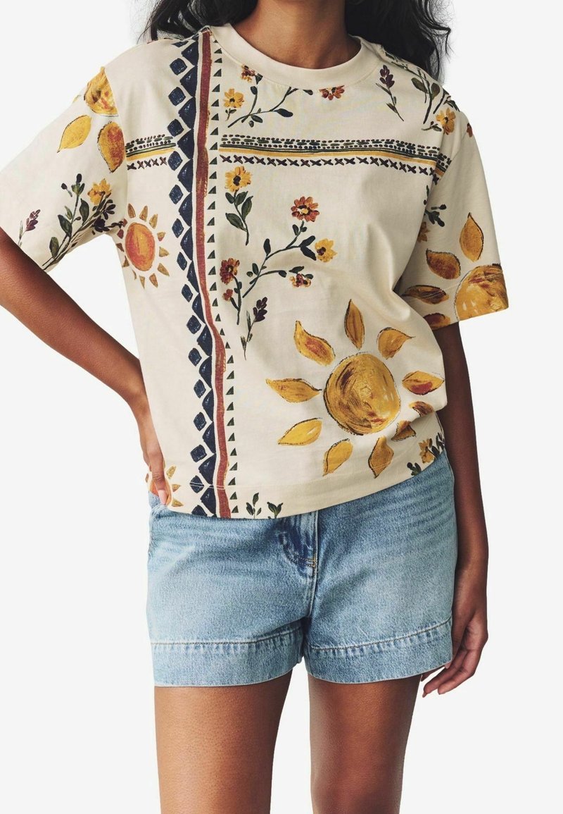 Femme portant un t-shirt beige à manches courtes avec des imprimés de tournesols et de fleurs, associé à un short en jean bleu clair.
