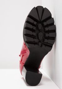 Czerwone lakierowane buty na czarnym gumowym podeszwie, z masywnym obcasem i wzorzystą przyczepnością. But posiada smukły design oraz szczegóły po bokach.