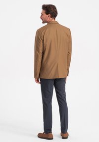 Ruskea bleiseri, jossa on rakenteellinen muotoilu, yhdistetty tummanharmaisiin housuihin. Bleiserissä on taitettu kaulus ja kaksi nappia, ja se on valmistettu sileästä kankaasta.