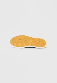 Podplat čevlja z teksturirano gumijasto površino, ki vsebuje krožni in valovit vzorec. Barva je pretežno rumena z belim robom.