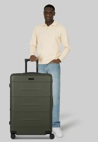 Maleta rígida verde con superficie texturizada, dos ruedas negras y un asa retráctil, situada junto a una persona con ropa casual.