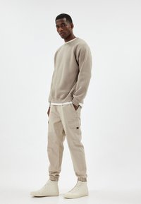 Beige sweatshirt med rund halsringning och insydda ärmar, kombinerad med ljusbeiga cargobyxor med sidofickor och elastiska muddars. Vita hög-top sneakers.