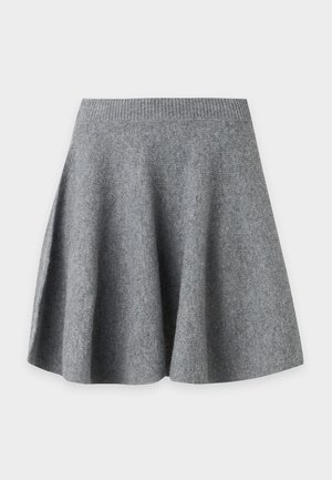 Kvinde iført en mørkegrå højhalset sweater, gråt plisseret skørt og sorte sandaletter med remme på en ensfarvet hvid baggrund.