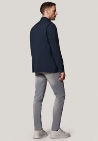 Granatowa wodoodporna kurtka o prostym kroju, z zapięciem na guziki przy mankietach i kieszeniami po bokach, w zestawie z jasnoszarymi dżinsami i neutralnymi trampkami.