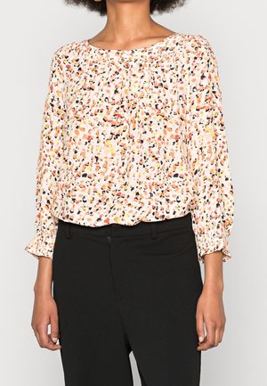 Kobieta w bluzce z długim rękawem w wielokolorowe abstrakcyjne plamy i czarnych spodniach, stojąca na jednolitym tle.