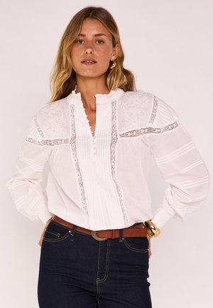 Donna con capelli biondi ondulati indossa una camicetta bianca di pizzo con pieghe, jeans scuri, cintura marrone, orecchini a cerchio dorati e braccialetto d'oro.